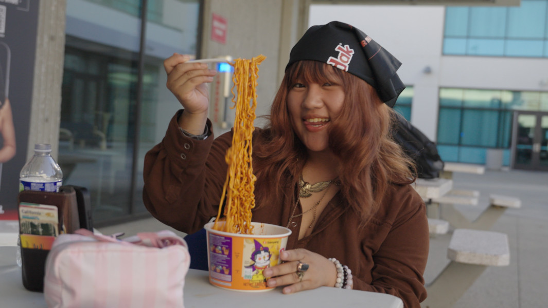 A girl eating a pot of instant noodles with her phone propped up filming a TikTok
